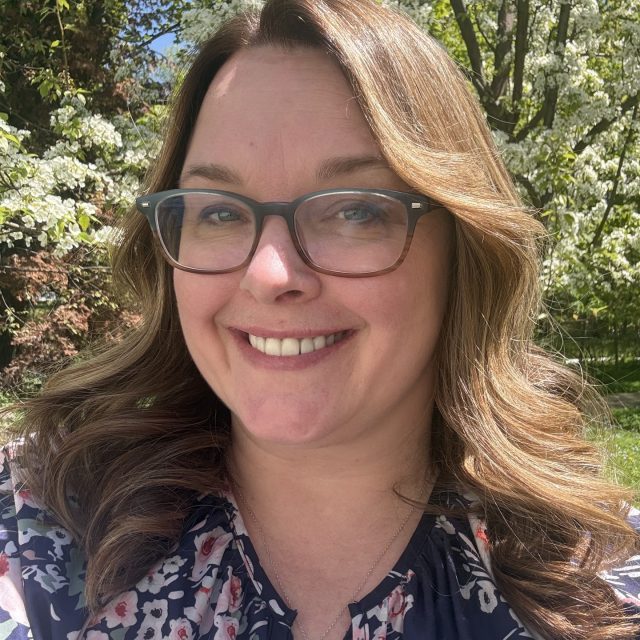 Woman with long brown hair and glasses with a blue and pink floral shirt.
